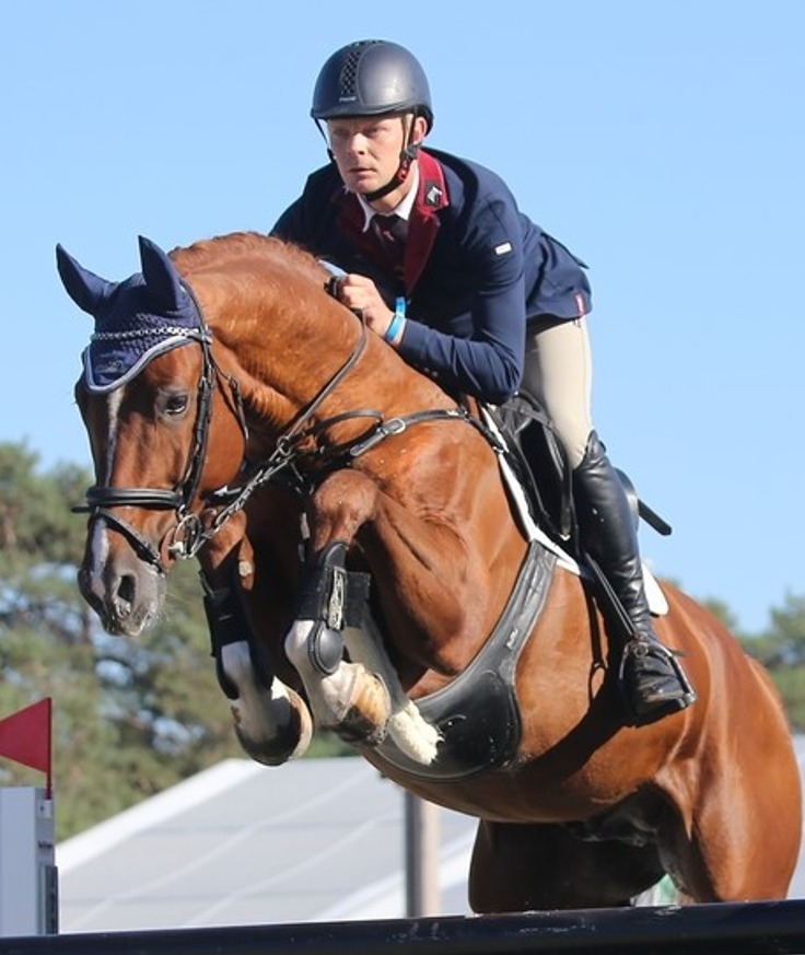 Jahač u natjecateljskoj uniformi na snažnom kestenjastom konju skače preko preponske prepreke pod vedrim nebom.
