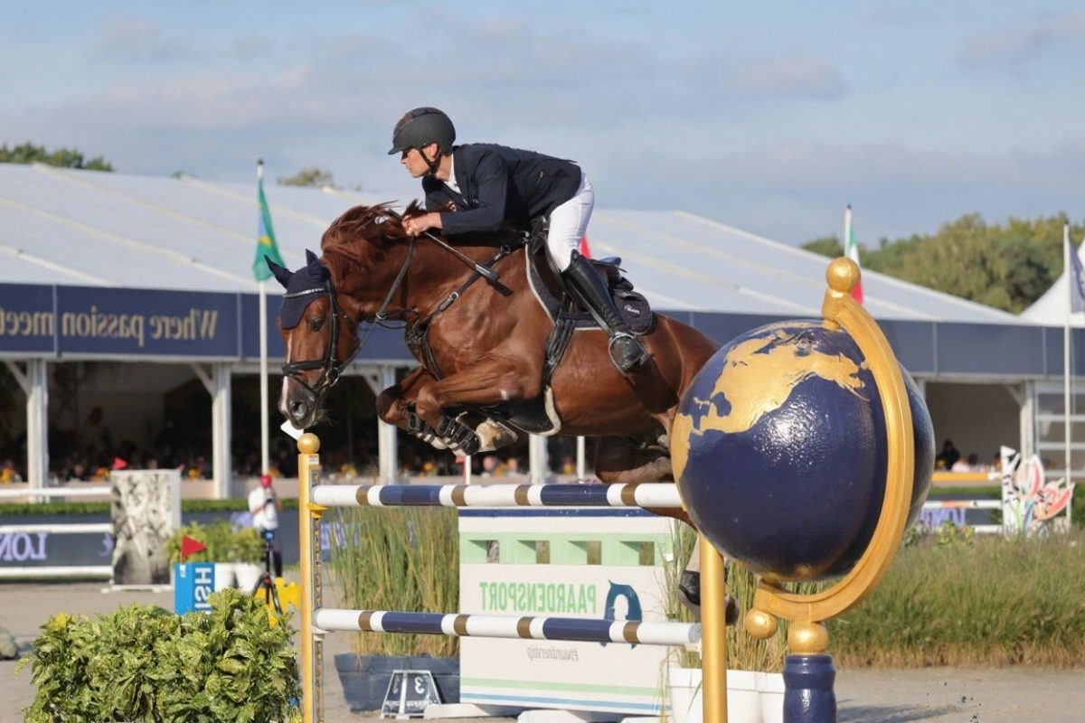 Ein Reiter auf einem braunen Pferd springt über ein hohes Hindernis mit einer Globusverzierung während eines Reitturniers im Freien, im Hintergrund sind Zuschauer und Zelte zu sehen.