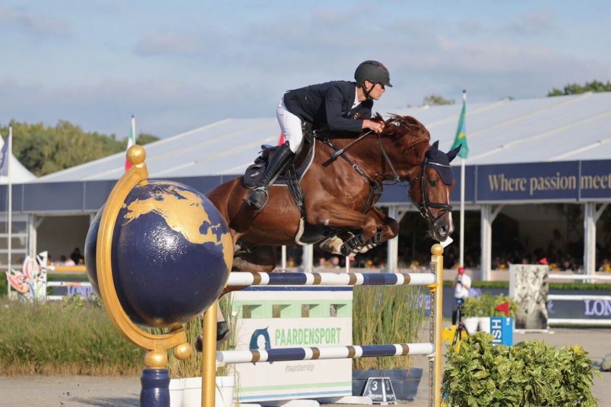 Ein Reiter auf einem braunen Pferd springt über ein hohes Hindernis mit einer Globusverzierung während eines Reitturniers im Freien, im Hintergrund sind Zuschauer und Zelte zu sehen.