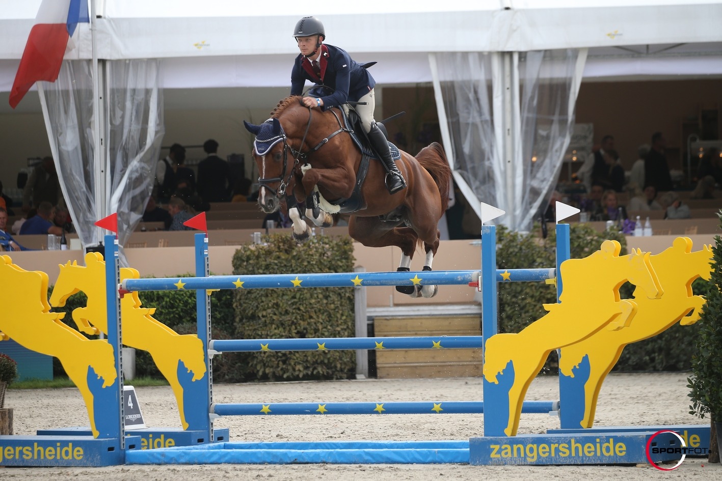 Jahač na konju skače preko prepoznatljive žuto-plave prepreke s motivom konja na turniru Zangersheide, pred gledateljima pod bijelim šatorom.