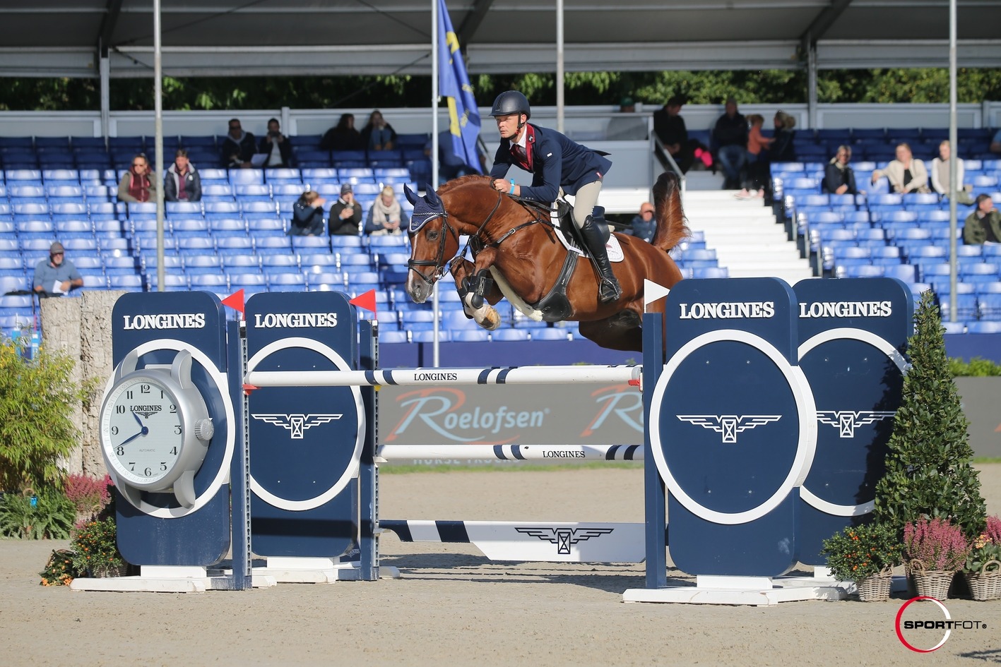 Jahač na smeđem konju u skoku preko Longines prepreke na preponskom turniru pred plavim tribinama s publikom.