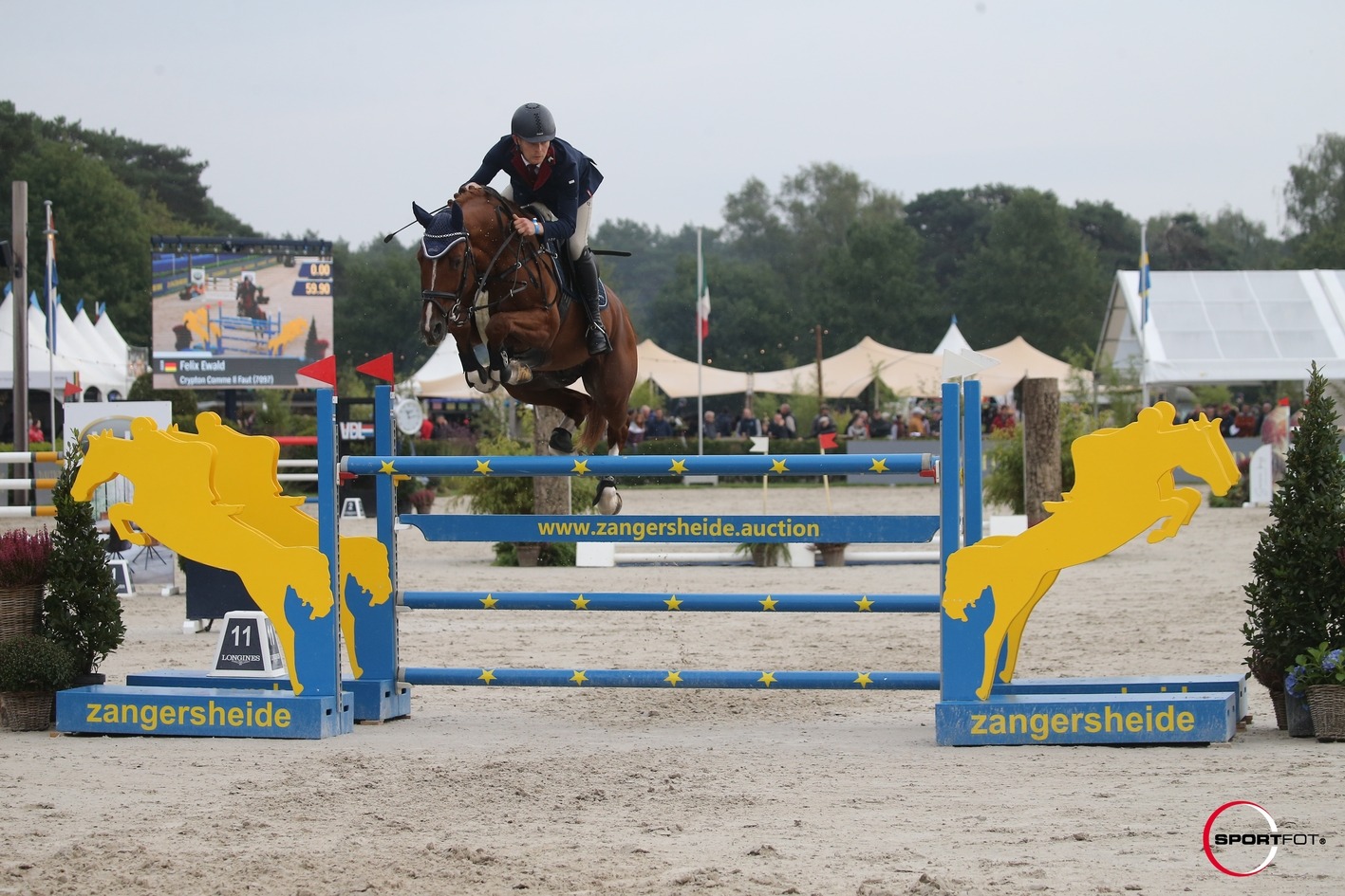 Felix Ewald na konju Crypton Comme Il Faut u skoku preko žuto‑plave Zangersheide prepreke na otvorenom natjecanju, s publikom i semaforom u pozadini.
