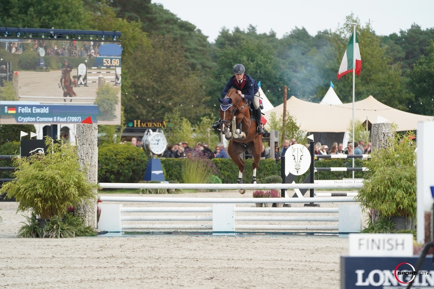 Jahač Felix Ewald na konju Crypton Comme Il Faut skače preko bijele prepreke kod cilja na otvorenom preponskom natjecanju, s gledateljima, zastavama i velikim elektronskim semaforom u pozadini.