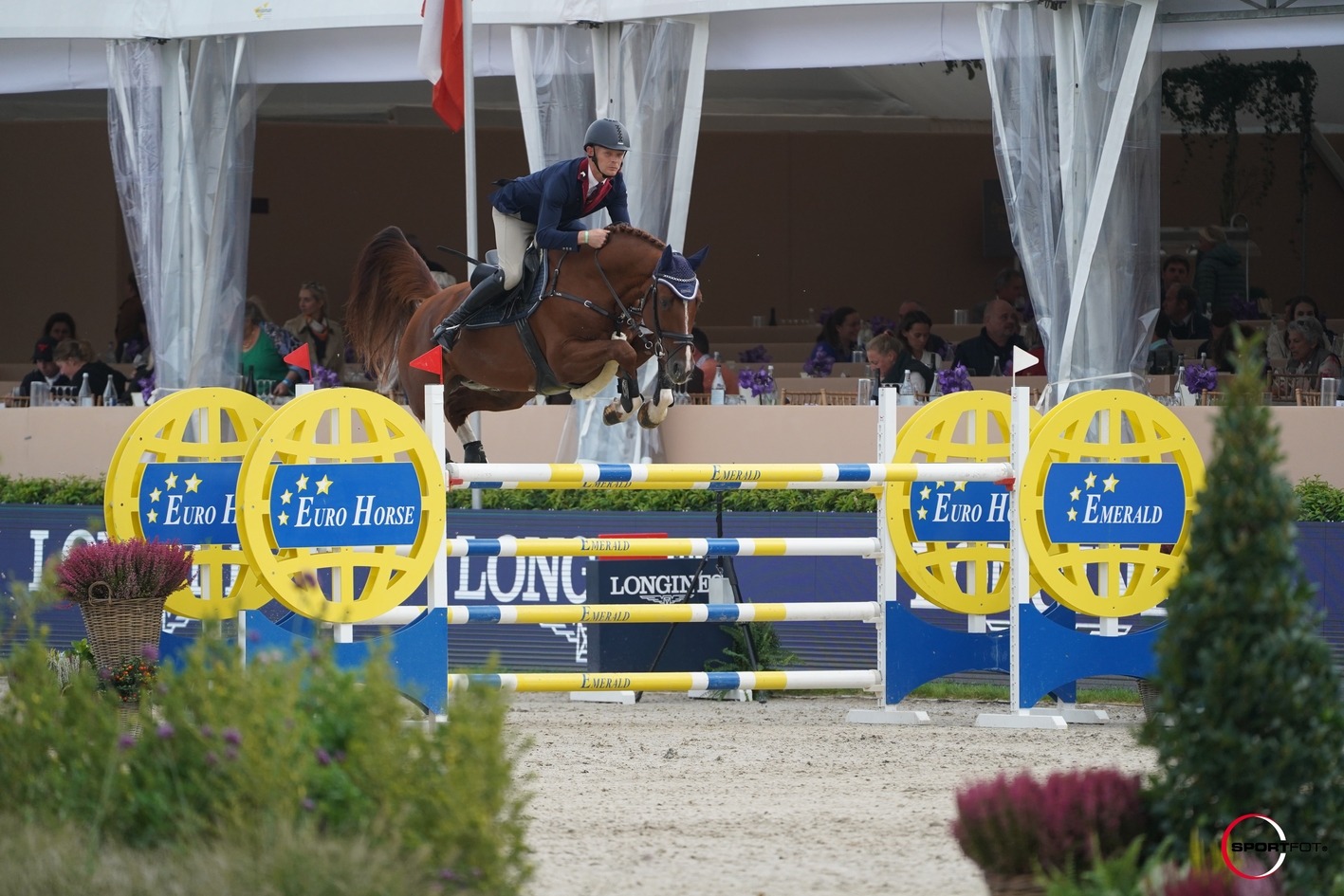 Jahač na smeđem konju u skoku preko žuto-plave prepreke Euro Horse Emerald, ispred publike pod bijelim šatorom na međunarodnom turniru.