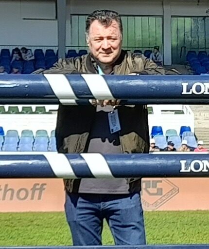 Muškarac stoji iza Longines prepona na konjičkom stadionu s praznim plavim i bijelim tribinama u pozadini.