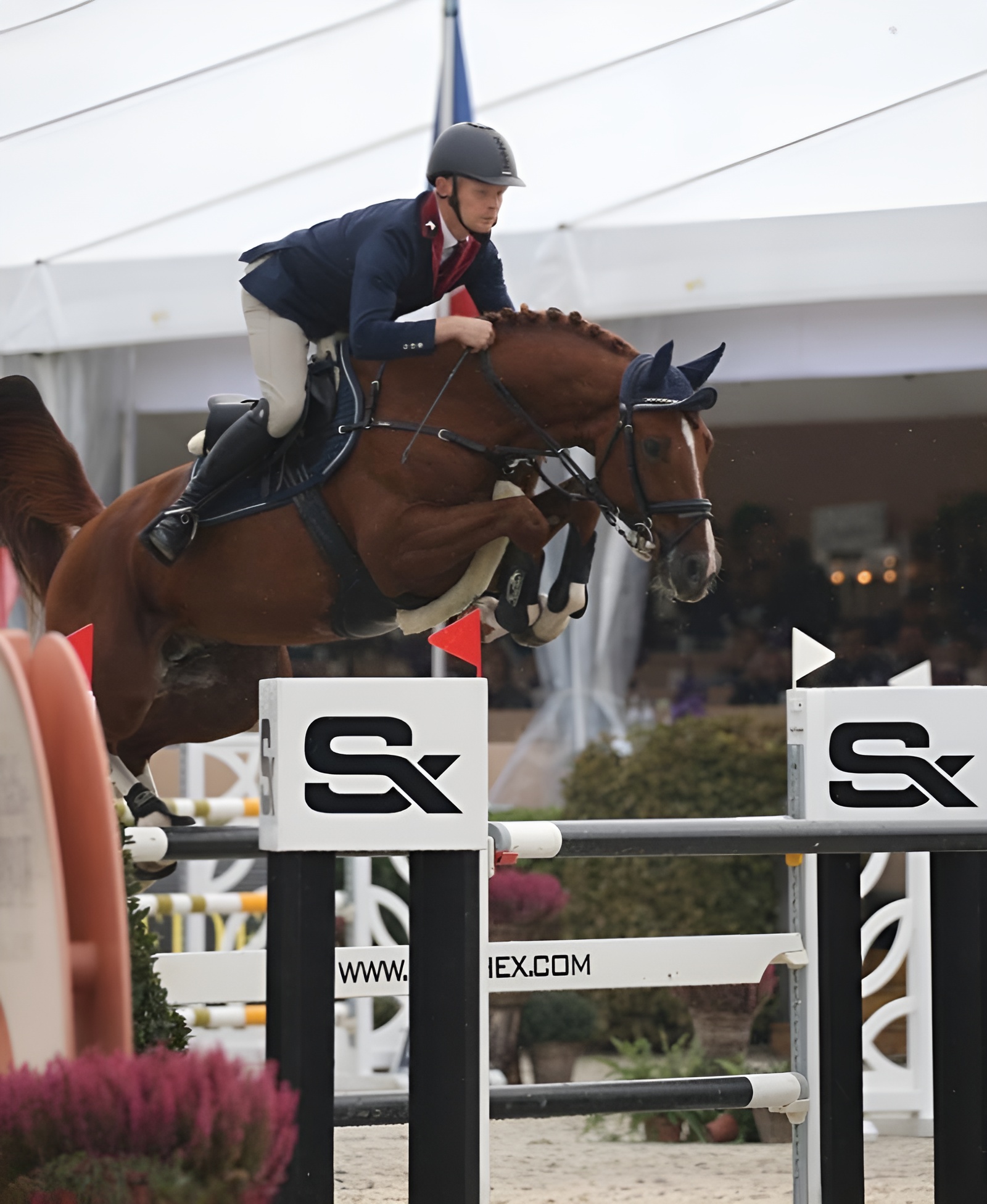 Jahač u natjecateljskoj uniformi i kacigi skače s konjem preko SX prepreke na preponskom turniru u zatvorenoj areni.
